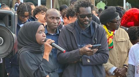Khalifa wearing a navy shirt and navy jacket, standing next to a crowd of people.