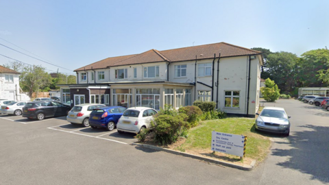 A small hospital. The building is white and has a brown roof. There are six cars in a car park surrounding it. The sky is blue