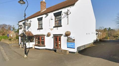 The outside of a white village pub with bay windows on the ground floor and a car park. The paintwork is chipped and it looks a bit lifeless.