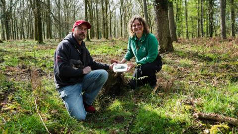 John Smith and Leoni Dawson crouch on the ground in the Forest of Dean holding the axe head between them in a plastic tub. Smith wears jeans, a grey hoody and a red baseball cap, and Dawson wears a green Forestry England sweater and dark trousers. 