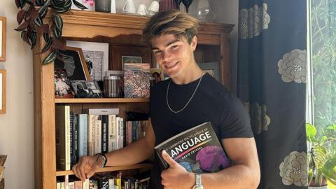 A young man with long brown hair, posing next to a bookcase full of books. He is wearing a black t-shirt and is holding a black book called "the Cambridge encyclopaedia of language"