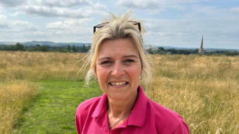 A blonde haired woman with glasses on her head. She is wearing a pink t-shirt with a necklace round her neck. There is a field and a blue sky with clouds in the background. 