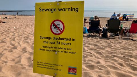 A beach scene with several people sitting on the sand facing the sea. In the foreground, a prominent yellow warning sign from East Devon District Council advises against bathing due to recent sewage discharge and warns to keep pets out of the water.