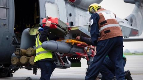 Two Royal Navy crew members in dark overalls work on a missile attached to a Wildcat/Lynx helicopter as it sits on the deck of a ship. The helicopter is dark grey
