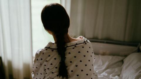 A girl with a long brown ponytail is wearing pyjamas with hearts on and sitting on a bed with her back to the camera. There are curtains in front of her.
