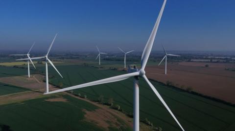 An aerial view of a wind farm in countryside. In the foreground is a very large white wind turbine. The tower supports three long blades. Several more wind turbines are arranged in across the fields. Low hedgerows separate some of the fields.