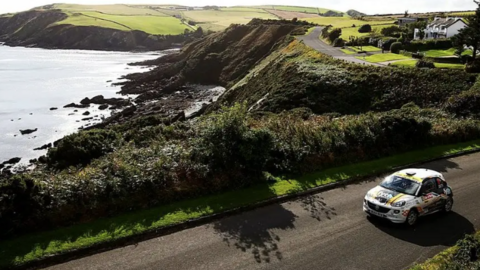 A rally car takes part in a previous event on the island, seen driving on a road near cliffs and sea