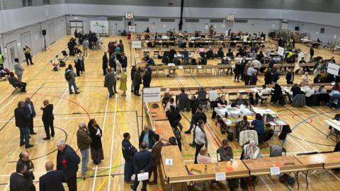 A sports hall with lots of tables in row and people milling around. Signs on the tables indicate geographical areas and identify this as an election count.