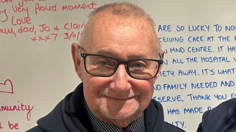 A man with very short hair, wearing black-rimmed glasses and with a short beard, smiles at the camera