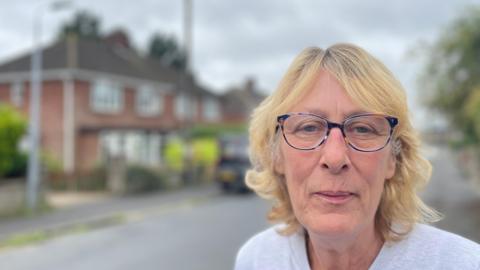 Lorraine Mitchell is wearing brown and black glasses, she has a blonde curly bob. She's standing in front of a house which has been blurred. 