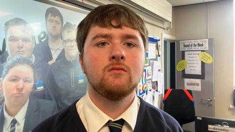 Ritchie has brown hair and a short beard. He's wearing a navy school jumper with a white shirt and a navy, blue and yellow striped tie. He's looking into the camera. He's standing in a classroom. 