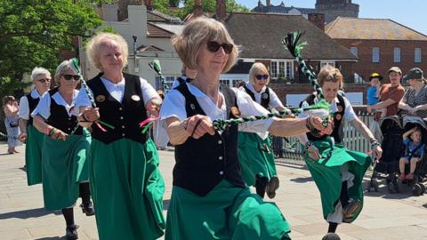 A troupe of eight clog dancers on a paved river bridge. They are wearing calf-length green skirts, black waistcoats and white, short-sleeved collared shirts. They are jigging and holding tassels. People are watching. 