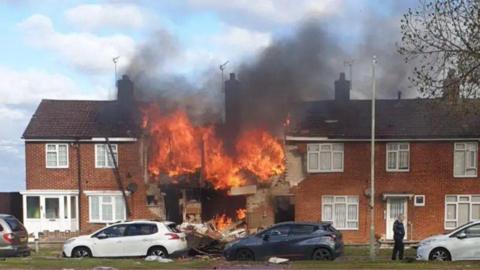 A row of terraced houses with cars parked along the road in front. The second house in from the left has been destroyed by an explosion and is completely on fire