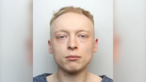 A mugshot of Miller, who has a vacant expression and is looking at the camera. He has short blonde hair and is wearing a blue top.