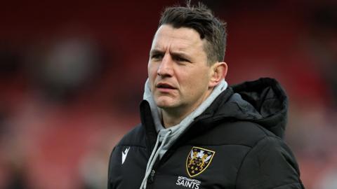 Northampton Saints director of rugby Phil Dowson, wearing a black coat with a grey hoodie underneath, watches his team from the touchline
