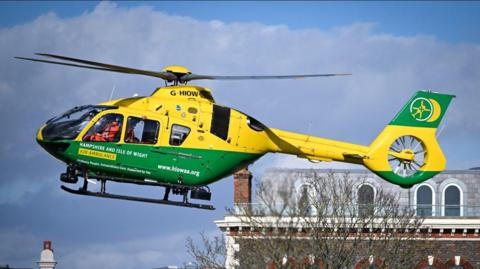 A yellow and green helicopter is pictured from the side, with its nose facing the left. It is in flight and a rooftop and the upper branches of a bare tree can be seen in the background. 