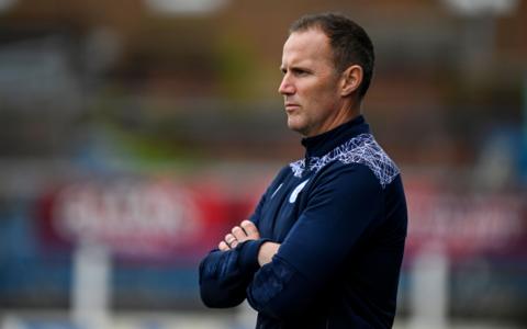 A football manager in a tracksuit top stands with his arms folded at the side of the pitch.