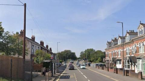 Wednesbury Road in Walsall has terraced houses on both sides. There is a bus stop and traffic on the road and trees in the background.