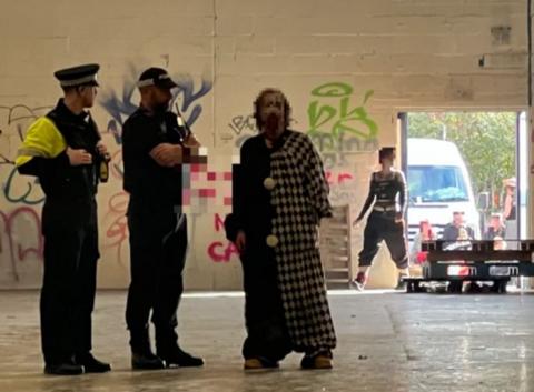 Two police officers inside a warehouse stood next to a man, whose face is pixilated. Graffiti is painted on the walls in the background