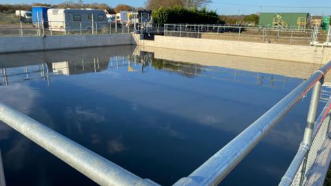 A large, square water talk, around the size of a square swimming pool, is surrounded on all sides by metal railings and a low brick wall. Industrial units can be seen in the background as well as a caravan.