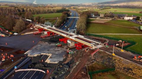 An aerial view of the new bridge being positioned over the M6. It is a light grey with bright red supports underneath rolling it into place.