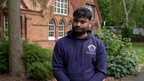 Sammey Ahmed is wearing a dark blue jumper with his name on, he's standing in front of a brick building with a tree next to him. 