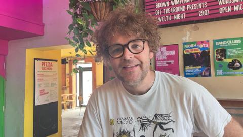 A man with short light brown curly hair and wearing a white T-shirt with a black and yellow design is smiling while sitting in the Future Yard music venue.