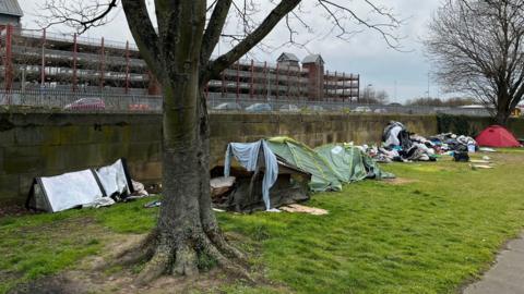 A number of tents are erected next to a concrete wall on a small strip of grass. There is a pile of bin bags and duvets on the grass between two of the tents. A multi-storey car park can be seen in the background.