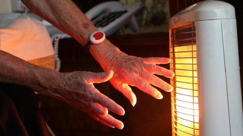 An elderly woman warms her hands at an electric fire
