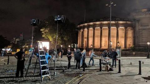 A film set at night with a crew setting up a scene on William Brown Street in Liverpool. Central Library is lit up in the background.