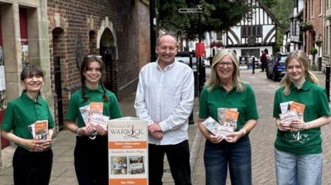 Four women in green T-shirts stand on a street holding leaflets. in the middle of them stands a man in a white shirt. In front of him is a sign advertising Warwick.