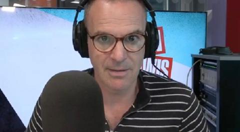 Martin Lewis wears black shirt with white stripes and glasses. He sits behind a microphone