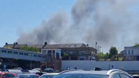 Smoke rising into the sky. The fire is not visible and appears to be taking place behind some buildings nearer the photographer. They are taking the image above cars in a car park.