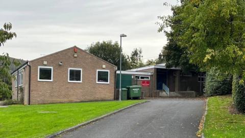 A school building. On the left is a building with a triangular roof and three windows. On the right is a building with a flat roof and small steps leading up to it. 