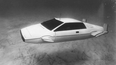The Lotus Esprit car featured in the 1977 film The Spy Who Loved Me. The white car, featuring protruding fins on its sides and and propellers on its back, is floating underwater near the sea bed, in this black and white image.