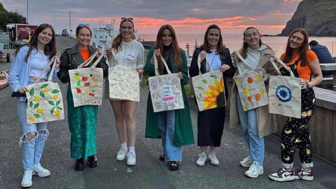 A group of seven girls hold up tote-bags with colourful designs. They are standing by the sea, with a headland and a pink sunset in the background.