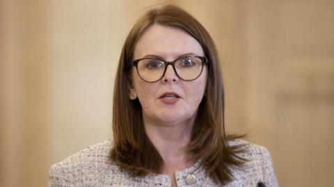 A woman with dark hair is wearing glasses and a grey jacket. She is in mid speech, and behind her is a beige wall. 