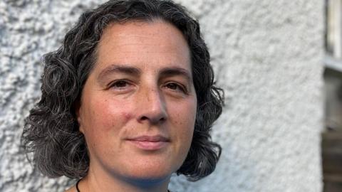 A woman with black and grey curly hair and brown eyes stands in front of a white wall