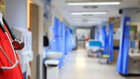 A hospital corridor, with booths either side screened by blue curtains. In the foreground is a male doctor wearing red scrubs and a stethoscope around his neck.