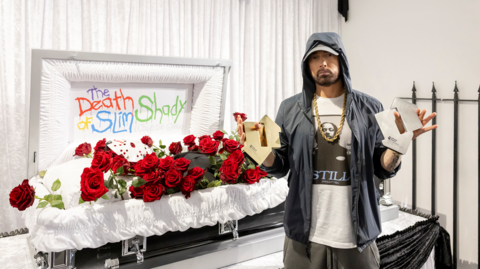 Eminem standing in front of a white coffin, holding two charts awards 