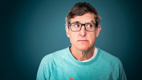 A headshot of Louis Theroux wearing a pake green jumper and glasses 