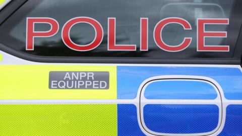 A zoomed up image of the side of a police car. There are yellow and blue reflective stickers, with a grey ANPR equipped sticker and a red sign saying 'Police' in the window.