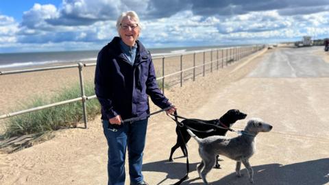 A woman with grey hair and glasses, walking a grey dog and a black dog on a lead, stands on the road at the edge of a beach. It is a sunny day but she is wearing a windproof coat, jeans and hiking boots. 