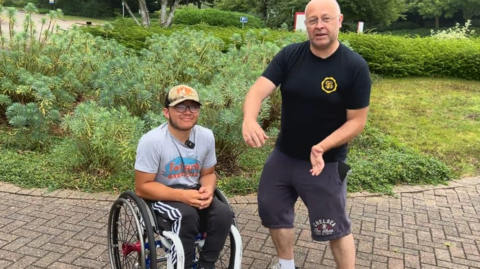 Ben in his wheelchair, wearing cap and a grey T-shirt and Adidas tracksuit bottoms, and his dad Michael next to him - wearing a black T-shirt and three-quarter length shorts with 'Chelsea - The Blues' on them - explaining something to a BBC Radio Berkshire journalist outside the BBC's offices in Reading