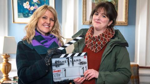 A woman with blonde hair and a woman with brown hair hold a clapper board between them which reads "SHETLAND". Ashley Jenson, the blonde woman, wears a dark green and purple scarf and a dark coat. Alison O'Donnell wears a patterned red scarf, a red jumper and green waterproof jacket. They are inside a formal looking room with baby blue wall panels and white outlines, with pictures hanging on the wall.
