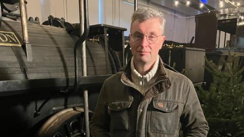 Neil Hammond, a man with glasses and short grey hair stands with his hands in the pockets of his jacket in front of a replica locomotive.