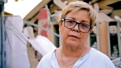 Zuly Marín, dressed in a white shirt and black glasses, stares into space.
