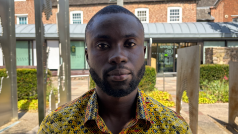 Noah Krah, a 26-year-old Ghanian student. He is wearing a bright yellow and orange button-up shirt. He has short black hair and a short beard. He has a stoic expression and looking directly at the camera.