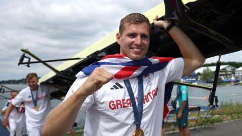 Rower Rory Gibbs carrying a boat on his shoulders with other members of the GB Eight at the Paris Olympics in 2024. He wears white GB kit and has a union flag around his shoulders and a gold medal hanging around his neck.