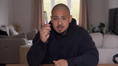 A man with a black hoodie is sat down at a desk or tablelooking straight into the camera. He is holding a 'pen' for diabetes in his right hand. In the background is are multiple white sofas, cream curtains, a tv and a houseplant
